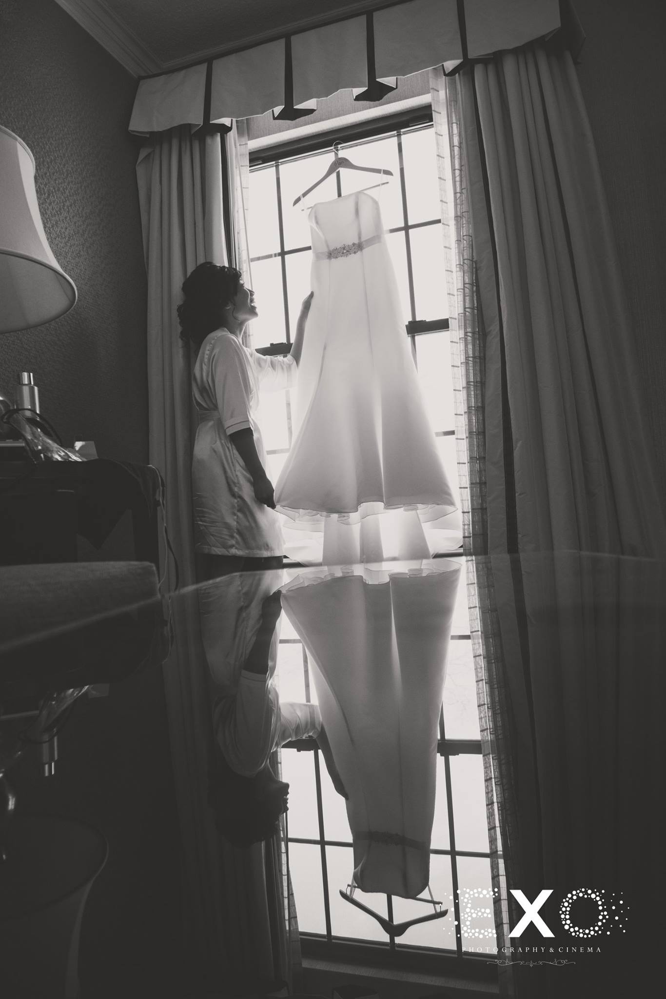 black and white image of bride with wedding gown hanging in window