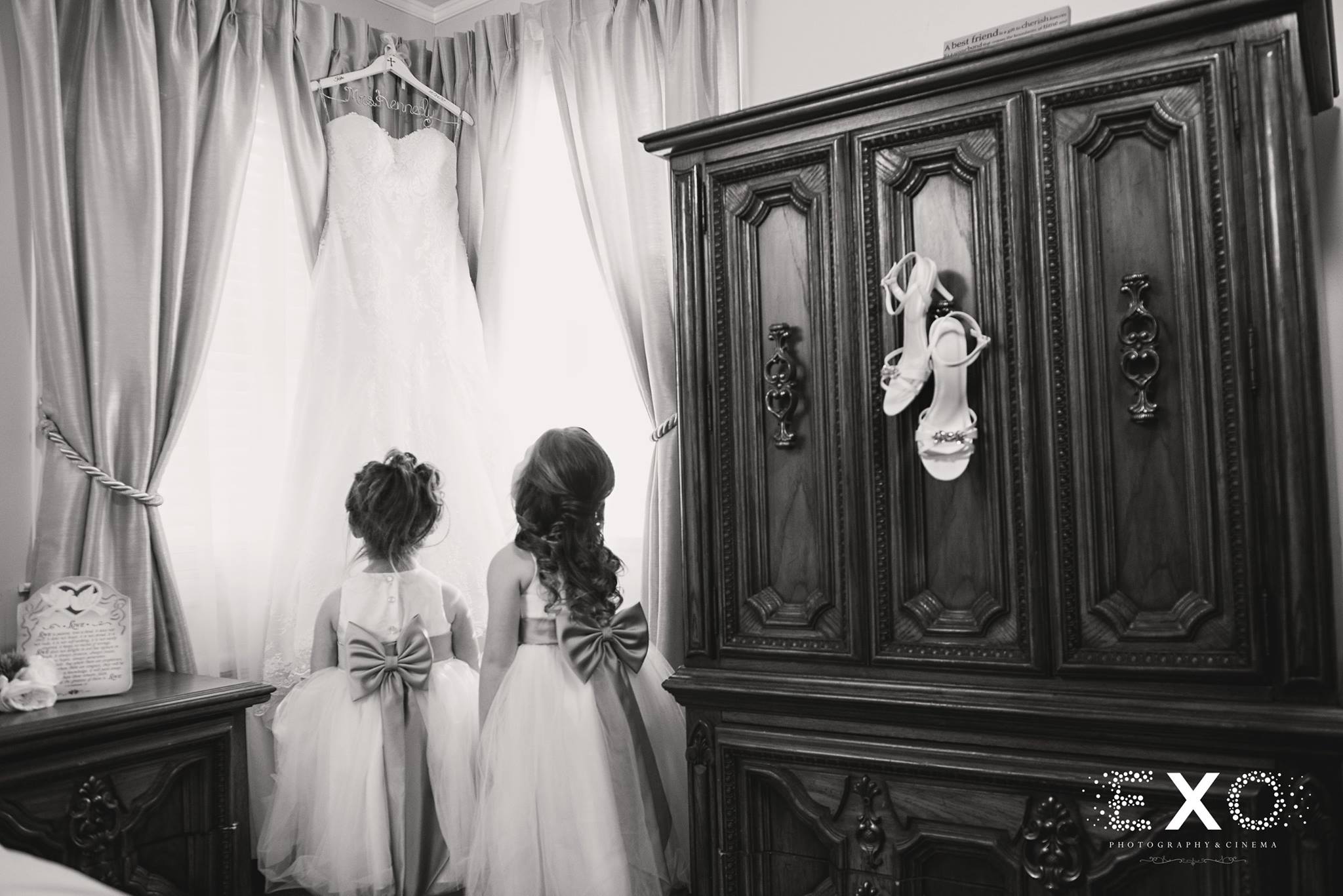 back and white image of flower girls looking at blossom bridal gown hanging in window at Heritage Club at Bethpage