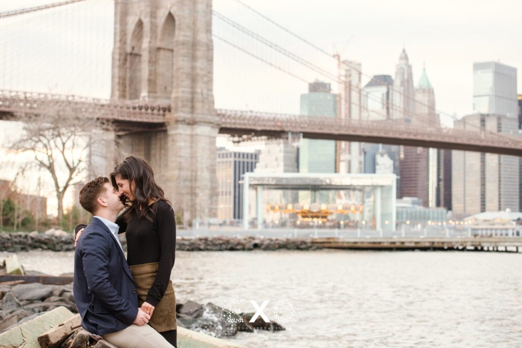 Couple looking at one another with bridge and carousel behind them at DUMBO