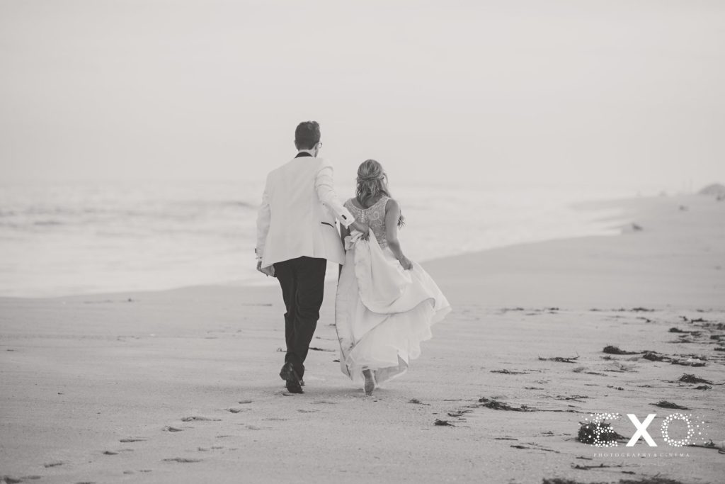 Groom holding brides dress while walking on the beach at Oceanbleu