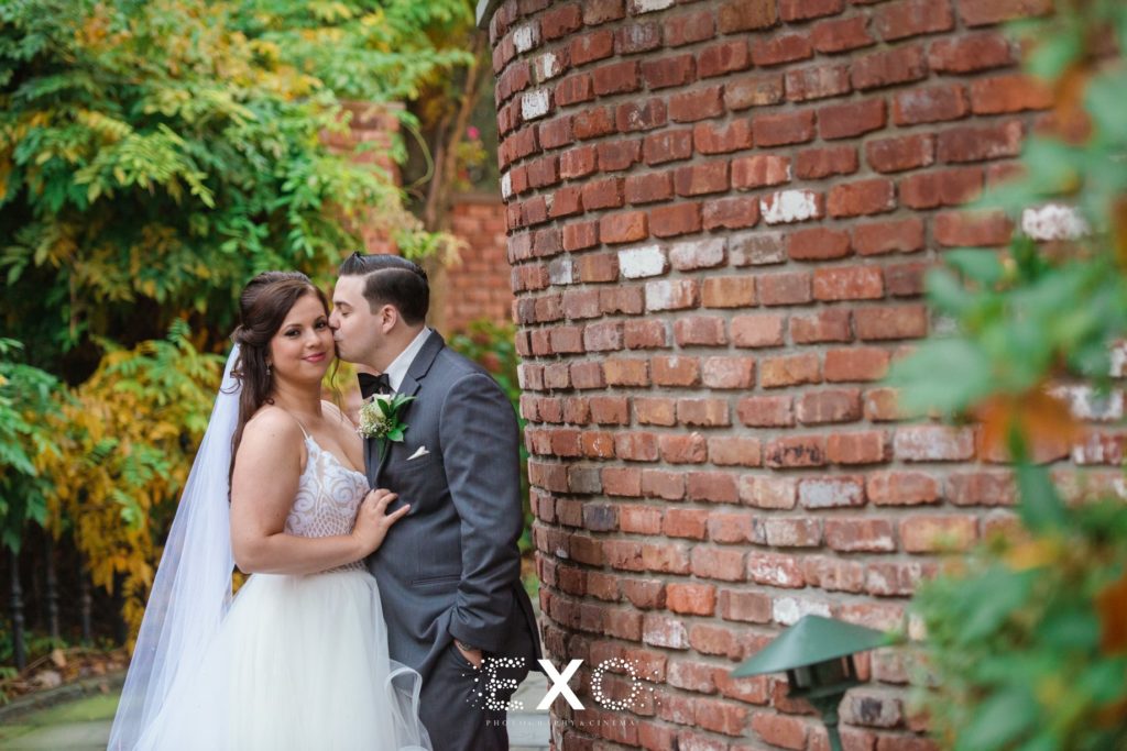 Groom kissing bride against brick wall at Fox Hollow