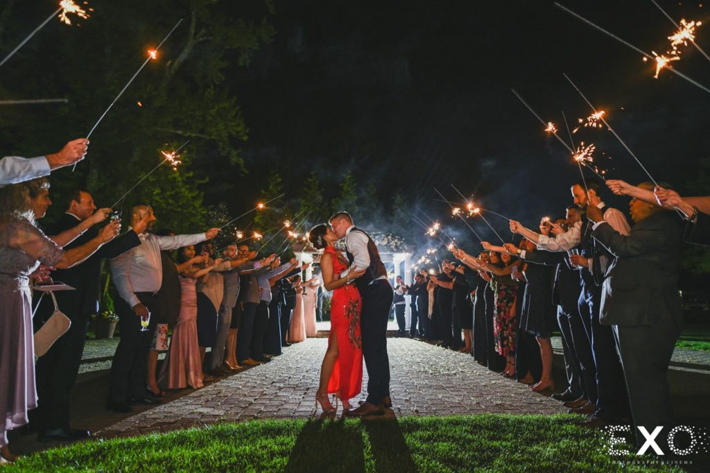 Bride and groom kiss outside surrounded by loved ones holding sparklers.