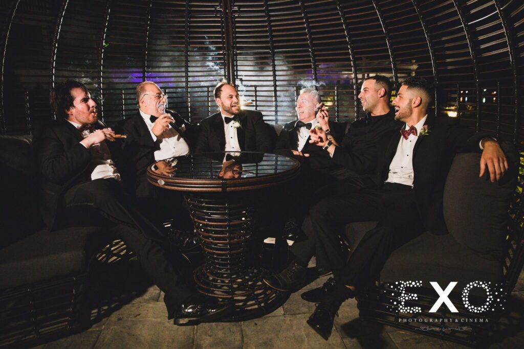Water's Edge wedding photo of groom and groomsmen smoking cigars by EXO Photography and Cinema