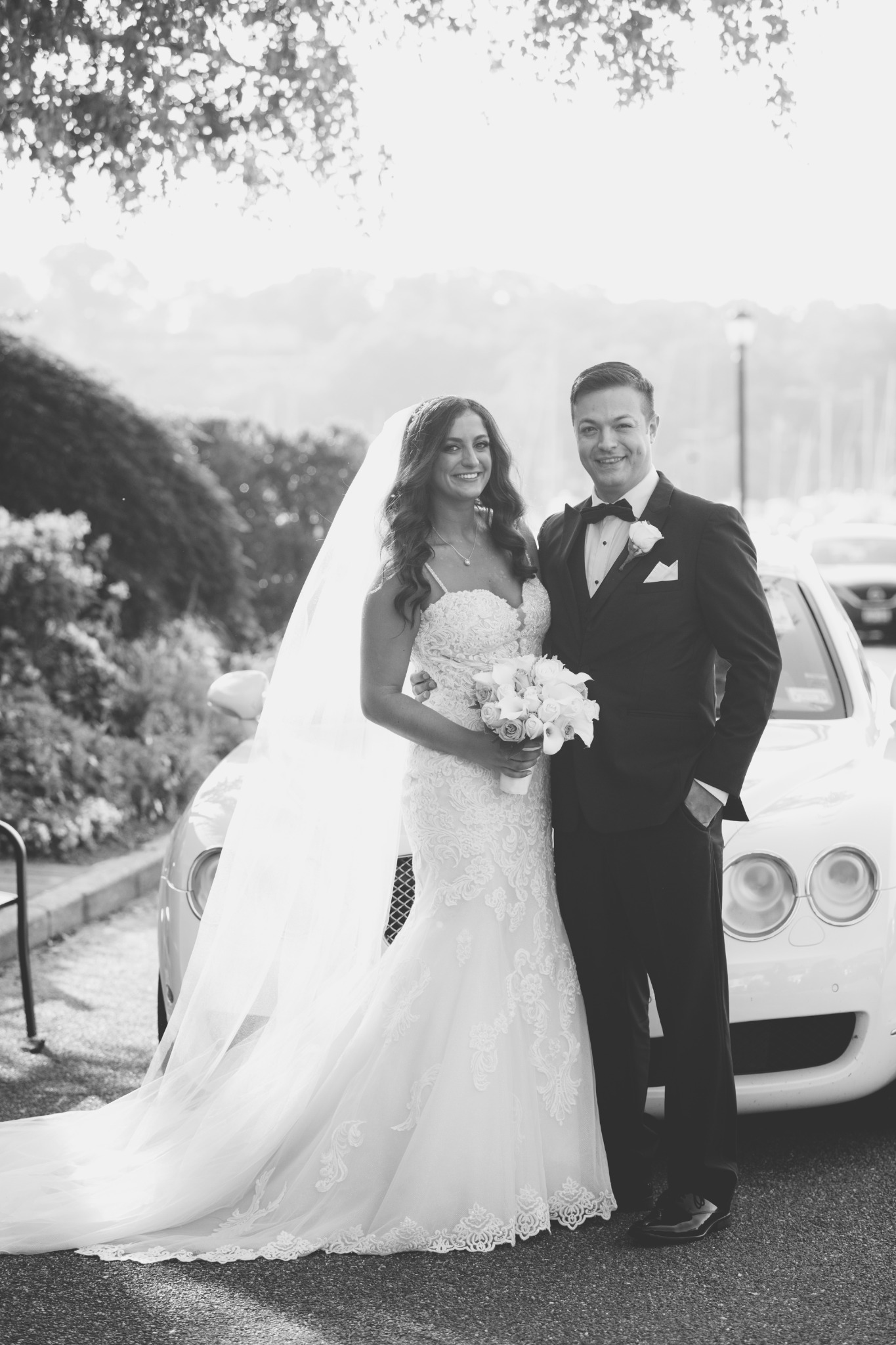 Black and white portrait of newlyweds beside a white car