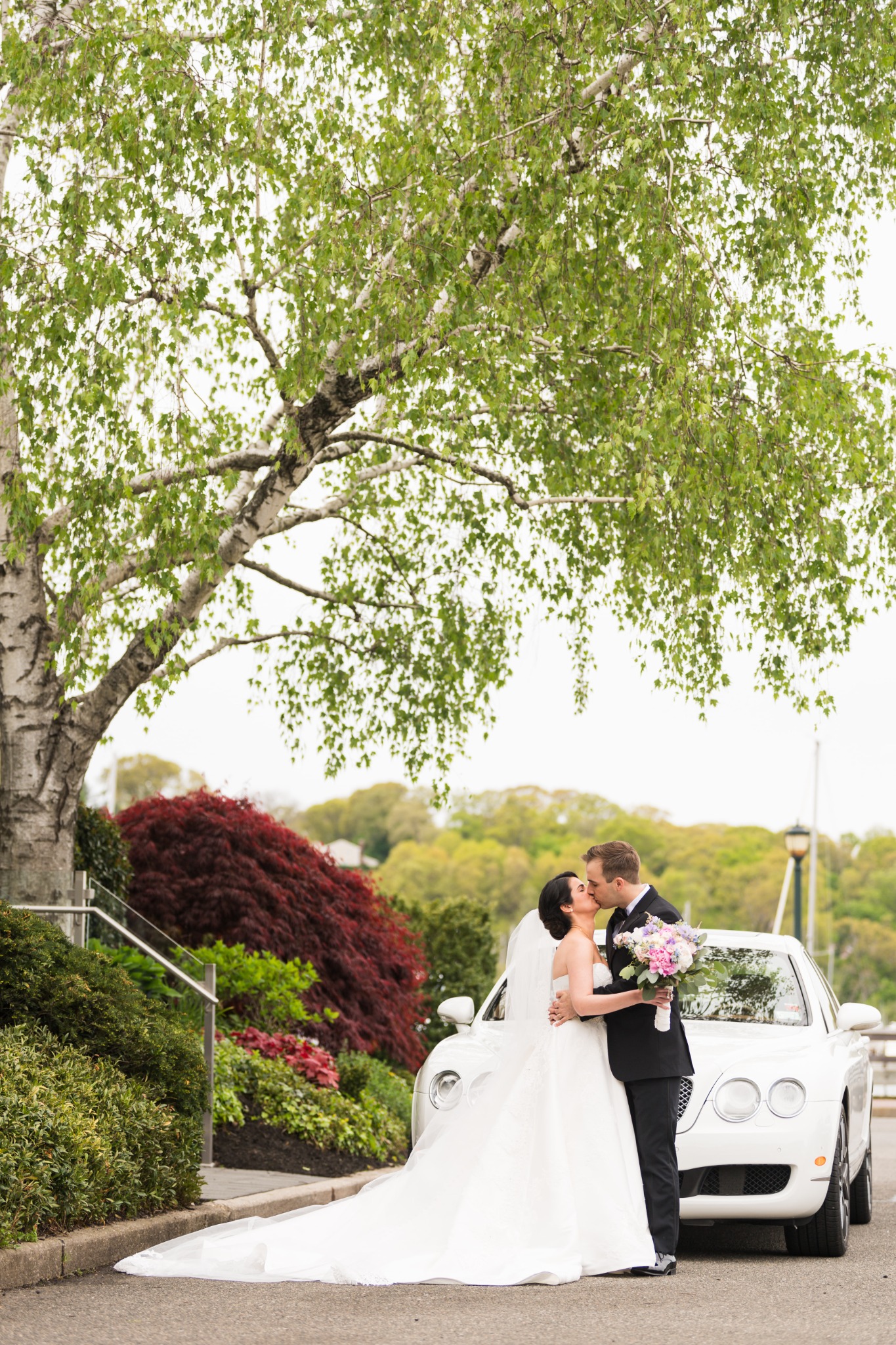 Bride and groom posing with a white car under a tree
