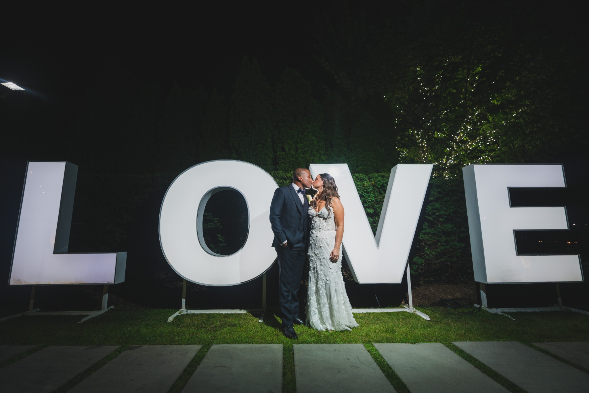 Newlyweds kissing beside illuminated LOVE letters at a nighttime wedding